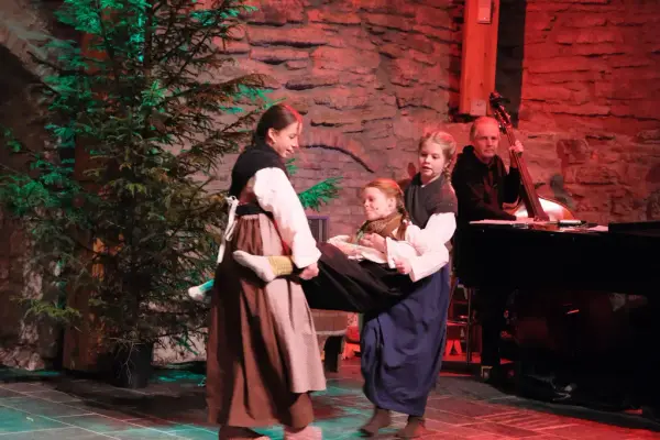 To jenter kledd i hverdagslige folkedrakter bærer en tredje jente i samme type klær mellom seg. Grantre i bakgrunnen.