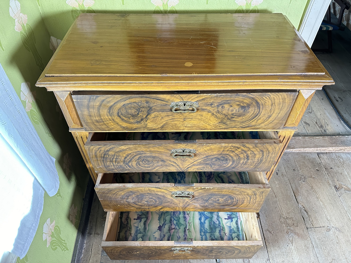 Byrå, av trä. Ådringsmålat i brunt. Profilerad kantig list upptill och nedtill. Svarvade fötter fram, fyrkantiga baktill. Fyra lådor. På ömse sidor av lådorna, fält med skurna prismor samt fält med kannelyrer. Beslag i gulmetall.