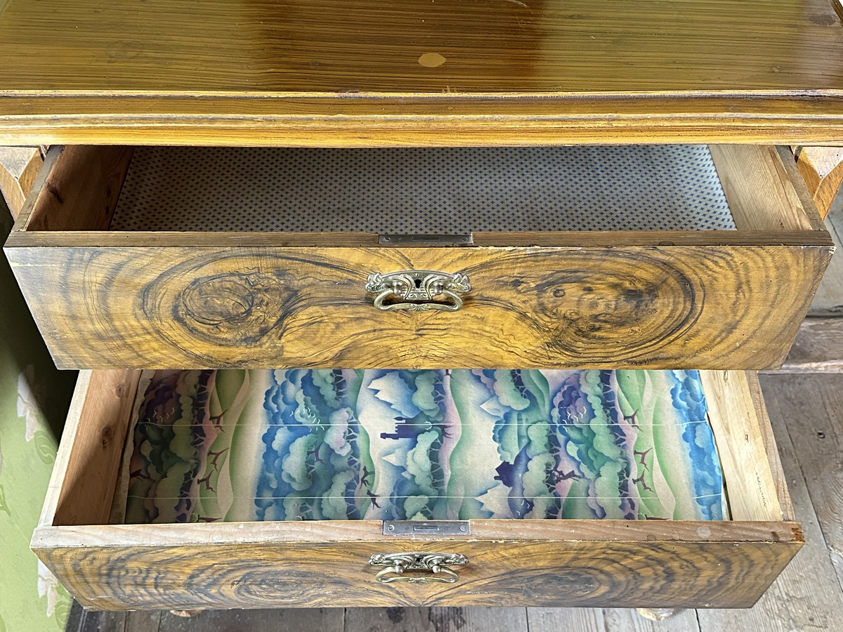 Byrå, av trä. Ådringsmålat i brunt. Profilerad kantig list upptill och nedtill. Svarvade fötter fram, fyrkantiga baktill. Fyra lådor. På ömse sidor av lådorna, fält med skurna prismor samt fält med kannelyrer. Beslag i gulmetall.