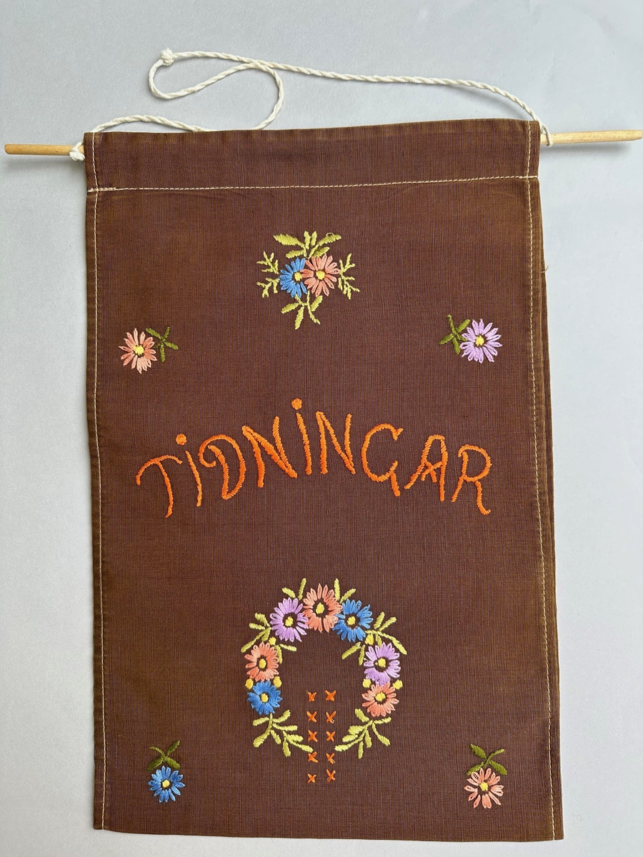 Tidningshållare brunt bomullstyg. Dubbelbvikt tyg som bildare en ögla i vilken tidningar hängs upp. Högst upp är en kanal sydd med en genomgående träpinne för upphängningen. Dekorerad med broderi i forma av blommor, en blomsterkrans och korsstygn. Broderat i både över mitten på hållaren: TIDNINGAR.