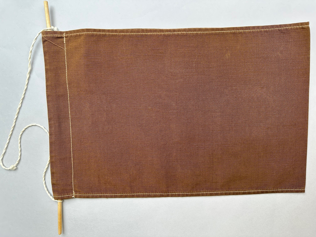 Tidningshållare brunt bomullstyg. Dubbelbvikt tyg som bildare en ögla i vilken tidningar hängs upp. Högst upp är en kanal sydd med en genomgående träpinne för upphängningen. Dekorerad med broderi i forma av blommor, en blomsterkrans och korsstygn. Broderat i både över mitten på hållaren: TIDNINGAR.