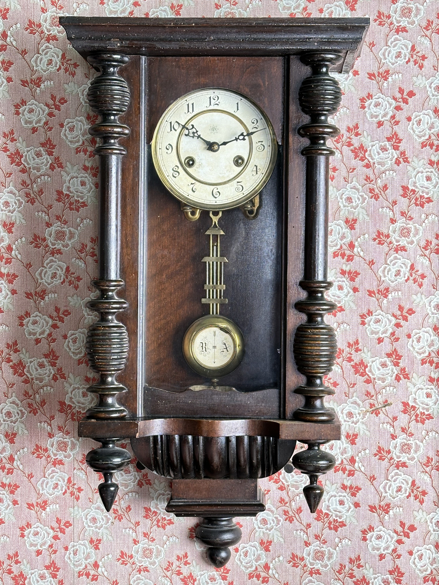 Väggur. Skulpterat fodral med svarvade ornament, av trä, målat i mörkbrunt. Vit urtavla med arabiska siffror, två hål med fyrkantiga piggar för att ställa visarna. Pendelverk. Öppningsbar framsida med glas. 