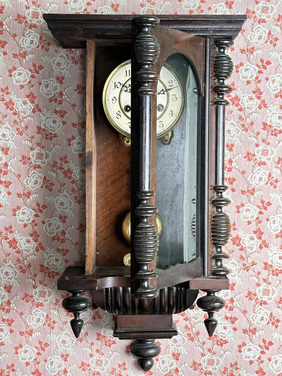 Väggur. Skulpterat fodral med svarvade ornament, av trä, målat i mörkbrunt. Vit urtavla med arabiska siffror, två hål med fyrkantiga piggar för att ställa visarna. Pendelverk. Öppningsbar framsida med glas. 