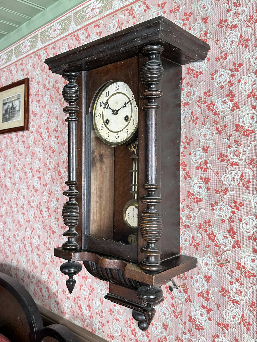 Väggur. Skulpterat fodral med svarvade ornament, av trä, målat i mörkbrunt. Vit urtavla med arabiska siffror, två hål med fyrkantiga piggar för att ställa visarna. Pendelverk. Öppningsbar framsida med glas. 