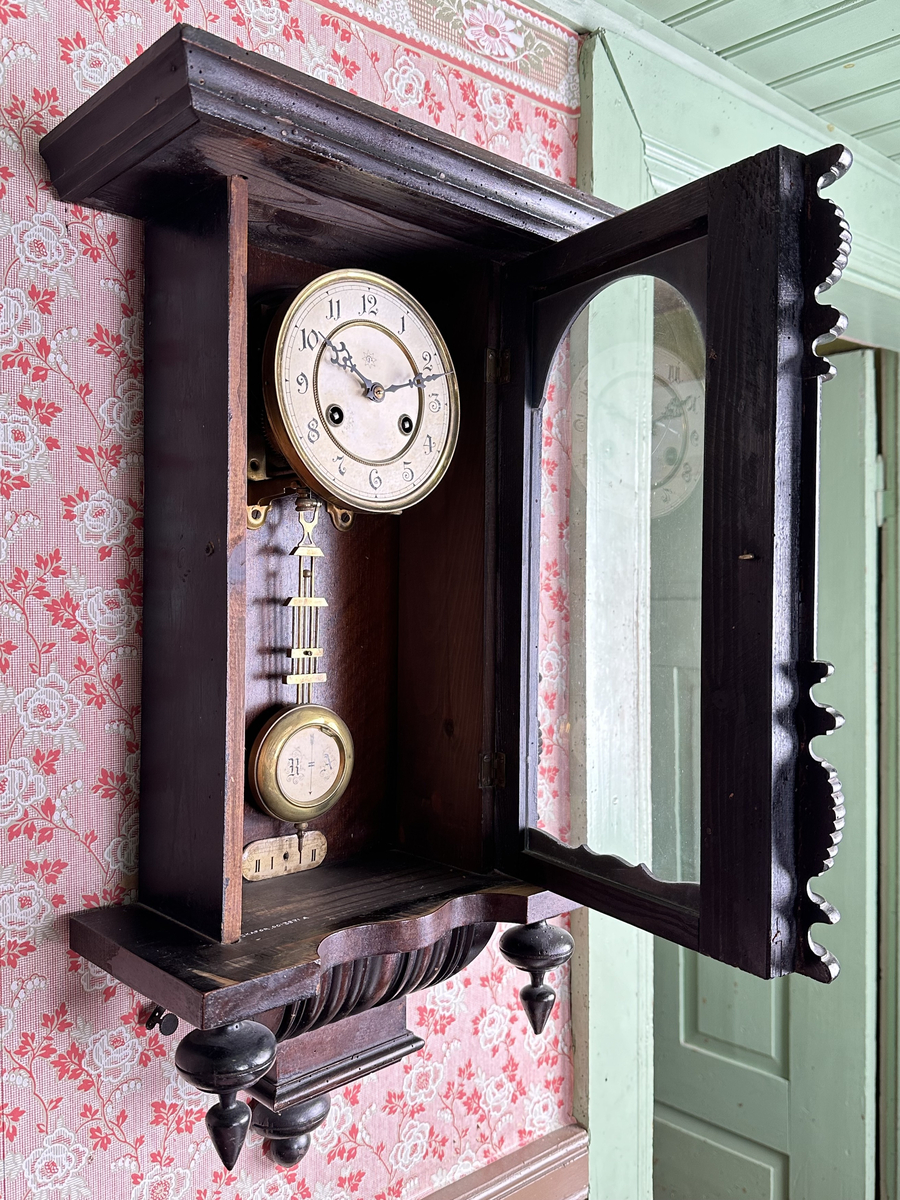 Väggur. Skulpterat fodral med svarvade ornament, av trä, målat i mörkbrunt. Vit urtavla med arabiska siffror, två hål med fyrkantiga piggar för att ställa visarna. Pendelverk. Öppningsbar framsida med glas. 