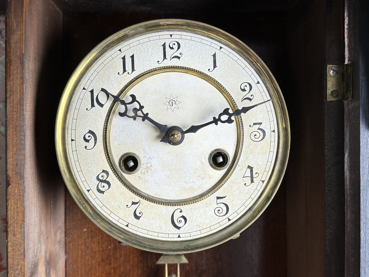 Väggur. Skulpterat fodral med svarvade ornament, av trä, målat i mörkbrunt. Vit urtavla med arabiska siffror, två hål med fyrkantiga piggar för att ställa visarna. Pendelverk. Öppningsbar framsida med glas. 