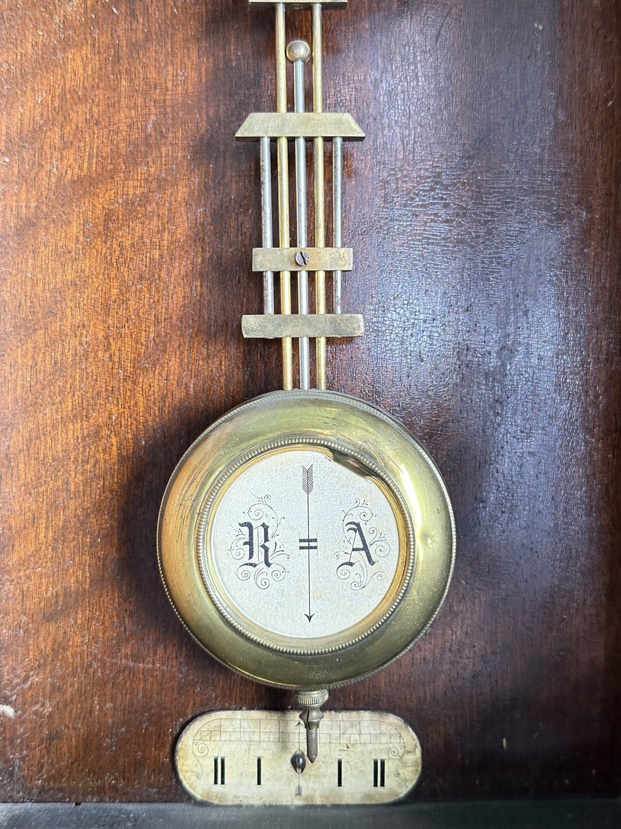 Väggur. Skulpterat fodral med svarvade ornament, av trä, målat i mörkbrunt. Vit urtavla med arabiska siffror, två hål med fyrkantiga piggar för att ställa visarna. Pendelverk. Öppningsbar framsida med glas. 