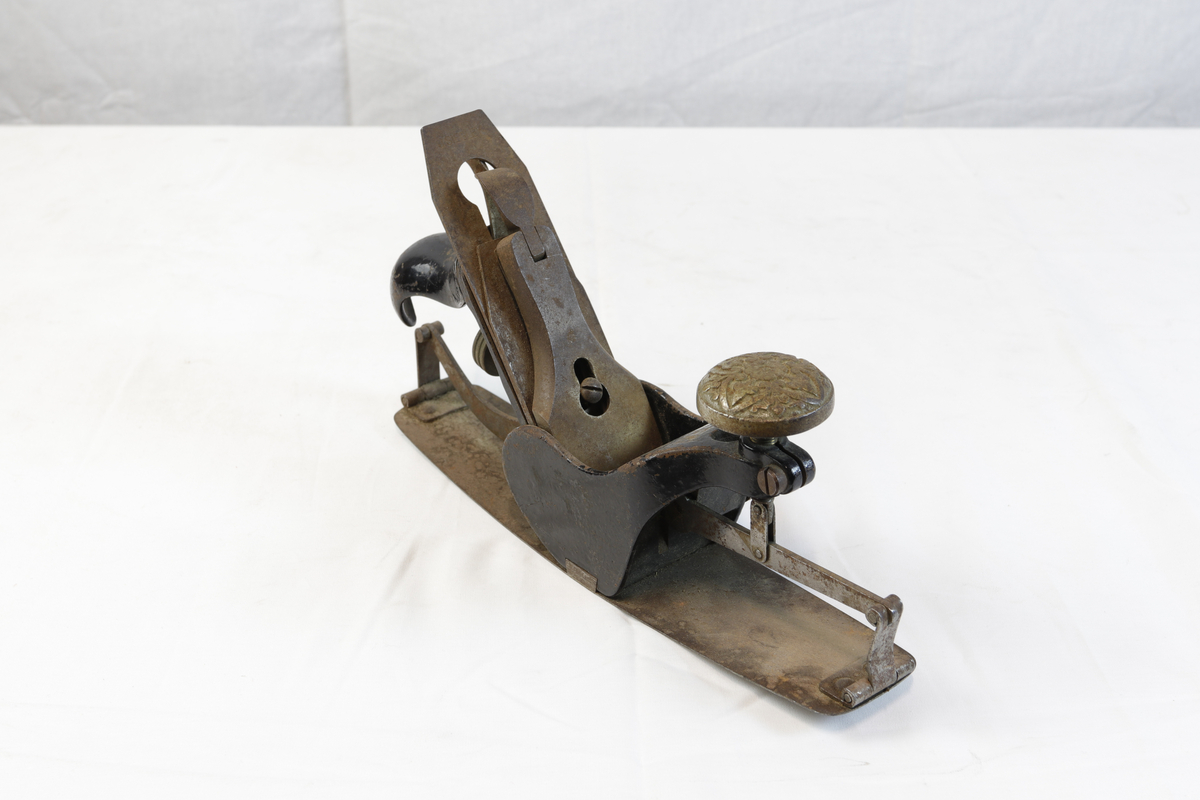 Justerbar buehøvel i metall. Håndtaket foran er dekorert med blomsterornamentikk. 
