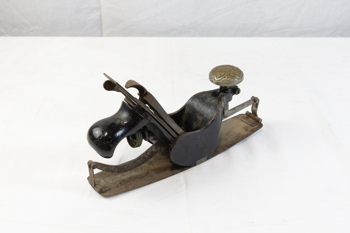 Justerbar buehøvel i metall. Håndtaket foran er dekorert med blomsterornamentikk. 
