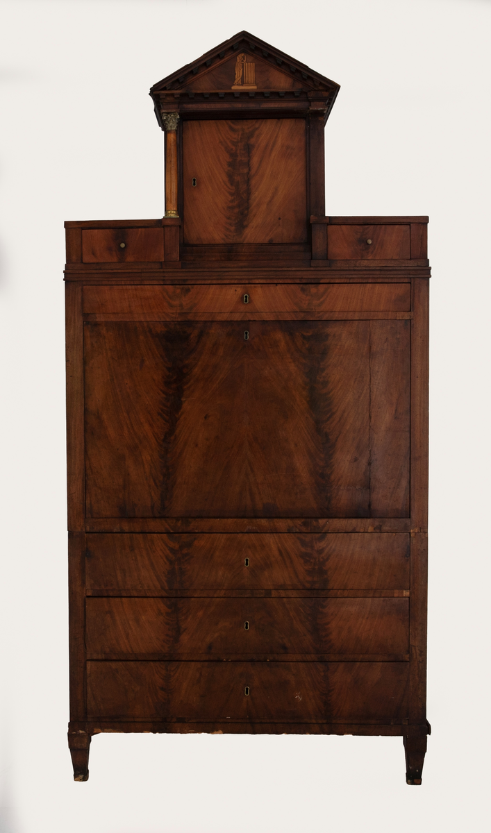 Skatoll i mahognifinér på tre. Nederst en skuffeseksjon med tre skuffer. Midtpartiet har skriveklaff, to skap og skuffer. Skapdørene viser intarsiamotiv forestillende kvinne ved søyle. På hver side av de to skapene er det marmorsøyler med forgylte bronsekapitler som dekker forsiden av smale, høye skuffer. Lave skuffer over og under skapene. Mellom skapene er det en åpning med speil og innlagt bunn. Øvre del av skatollen har et skap og to skuffer, en på hver side av skapet, med søyler av tre på hver side av skapdøren. Skatollen avsluttes med en gavl. Gavlfeltet har samme motiv som skapdørene i midtpartiet. Lave pyramideformete ben.