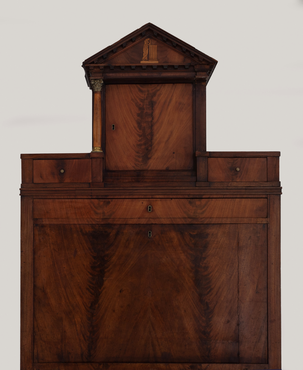 Skatoll i mahognifinér på tre. Nederst en skuffeseksjon med tre skuffer. Midtpartiet har skriveklaff, to skap og skuffer. Skapdørene viser intarsiamotiv forestillende kvinne ved søyle. På hver side av de to skapene er det marmorsøyler med forgylte bronsekapitler som dekker forsiden av smale, høye skuffer. Lave skuffer over og under skapene. Mellom skapene er det en åpning med speil og innlagt bunn. Øvre del av skatollen har et skap og to skuffer, en på hver side av skapet, med søyler av tre på hver side av skapdøren. Skatollen avsluttes med en gavl. Gavlfeltet har samme motiv som skapdørene i midtpartiet. Lave pyramideformete ben.