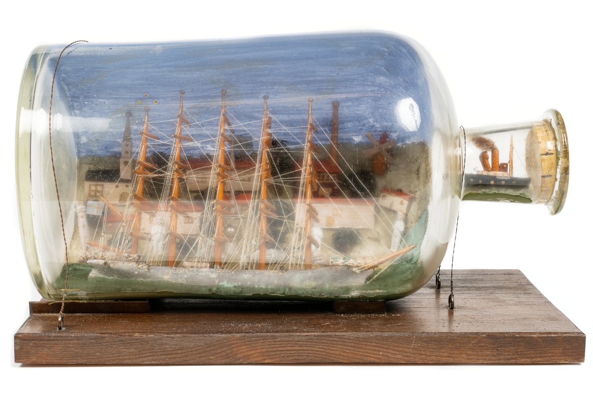 Flaskskepp med femmastad fullriggare. Seglen är beslagna och i flaskans hals är en liten bogserbåt behjälplig. I bakgrunden är en bebyggd strand. Tillverkaren är okänd.