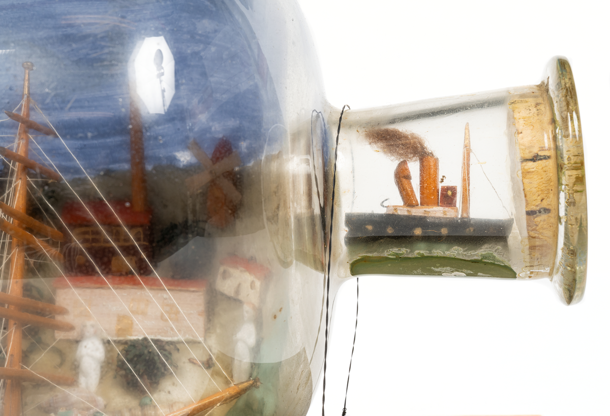 Flaskskepp med femmastad fullriggare. Seglen är beslagna och i flaskans hals är en liten bogserbåt behjälplig. I bakgrunden är en bebyggd strand. Tillverkaren är okänd.