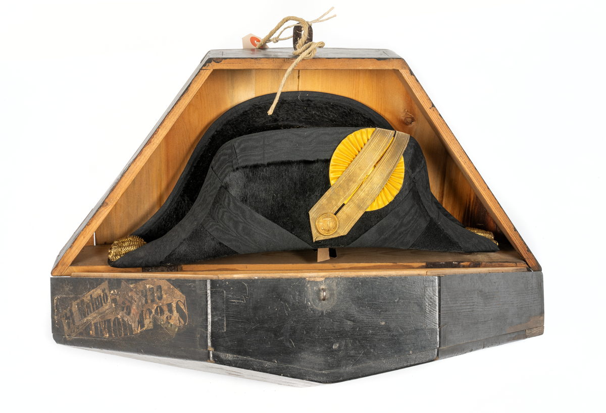 Paradhatt med fodral av trä. Hatten trekantig. Av snart silkesfelb med svarta kantband. Gul kokard med guldgalon och ankarknapp. Buljoner i ändarna. Rödd foder av siden. Firmastämpel (otydlig). Käkrem.