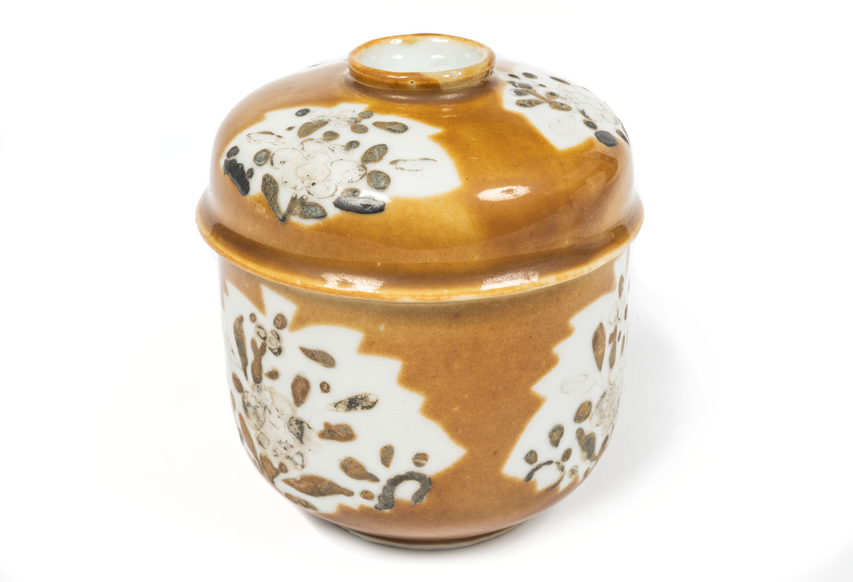 Bonbonier, bonbonjär. Skål med lock av ostindiskt porslin. Glaserad med brungul färg, vita bladformade fält med blomdekor i svart emaljfälrg. Enligt uppgift upptagen från den vid Hunnebådan sjunkna Ostindiefararens Göteborg".