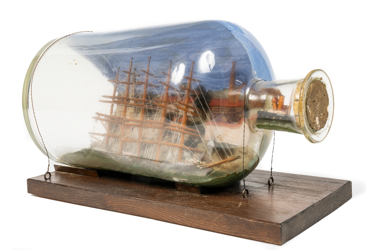 Flaskskepp med femmastad fullriggare. Seglen är beslagna och i flaskans hals är en liten bogserbåt behjälplig. I bakgrunden är en bebyggd strand. Tillverkaren är okänd.