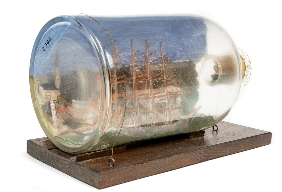 Flaskskepp med femmastad fullriggare. Seglen är beslagna och i flaskans hals är en liten bogserbåt behjälplig. I bakgrunden är en bebyggd strand. Tillverkaren är okänd.