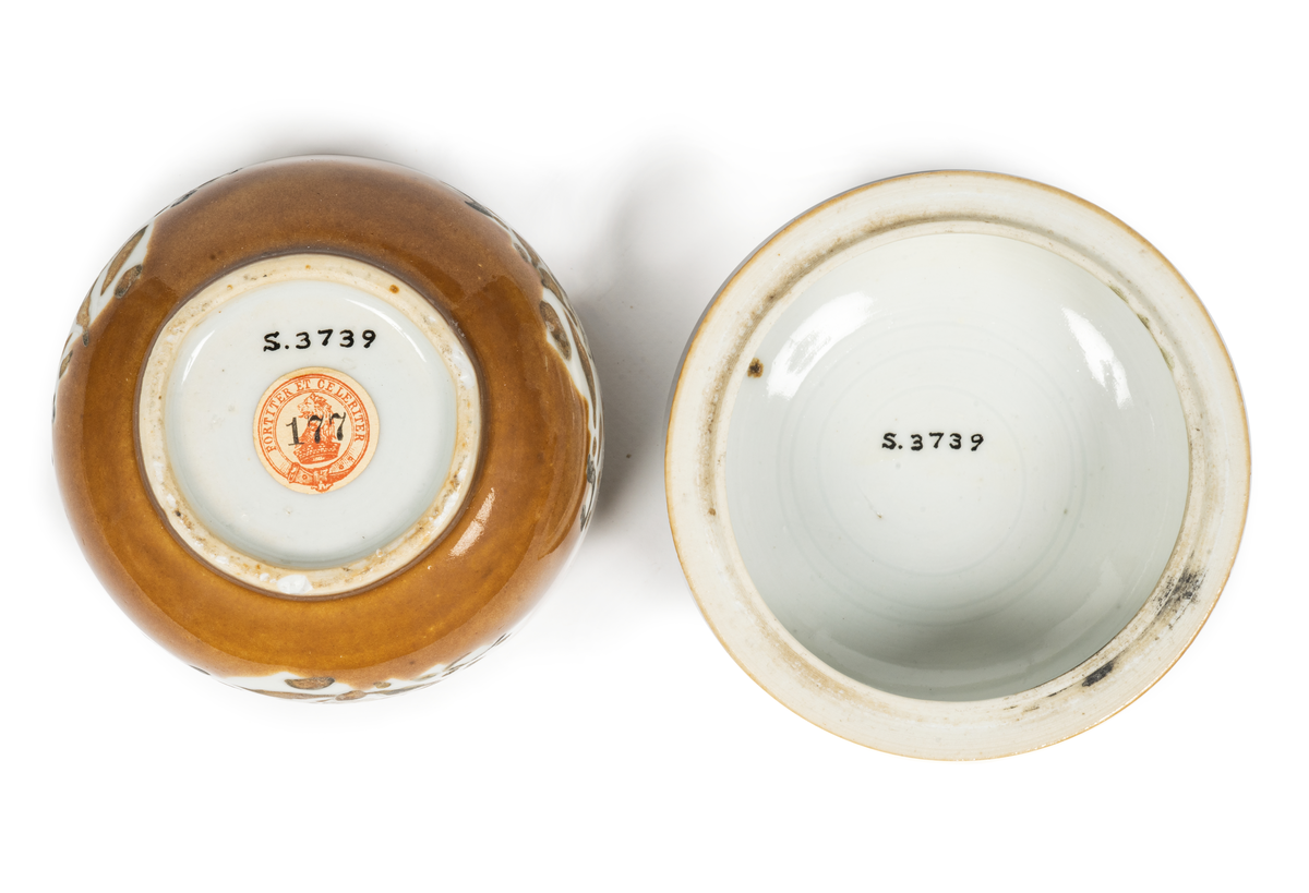 Bonbonier, bonbonjär. Skål med lock av ostindiskt porslin. Glaserad med brungul färg, vita bladformade fält med blomdekor i svart emaljfälrg. Enligt uppgift upptagen från den vid Hunnebådan sjunkna Ostindiefararens Göteborg".