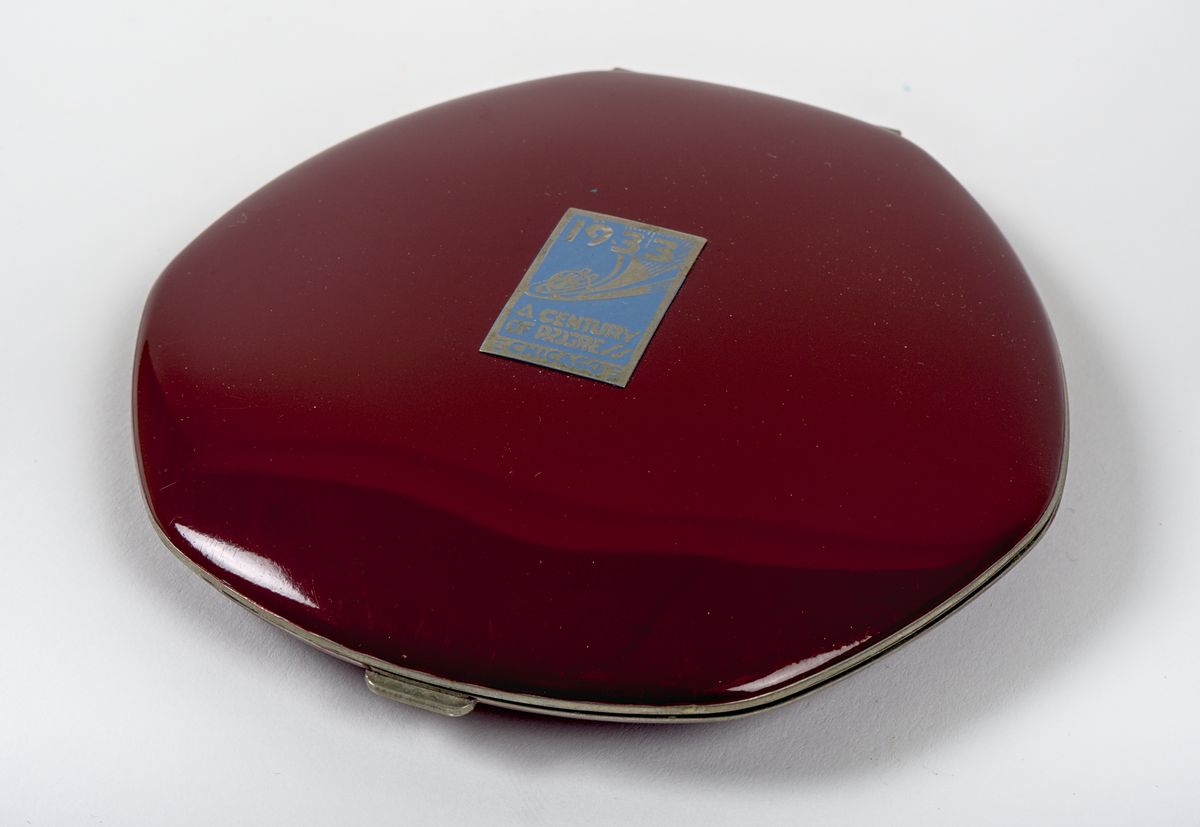 Oval puderdosa i metall med vinröd emaljerad yta. På ovansidan finns en metallplatta med silverfärgad text mot blå bakgrund: "1933, A Century of Progress, Chicago".

På insidan av locket sitter en spegel, medan botten har en rosa tyginsats för puder, märkt "ELGIN VANITY". Dosan stängs med en låsmekanism kopplad till en knapp.