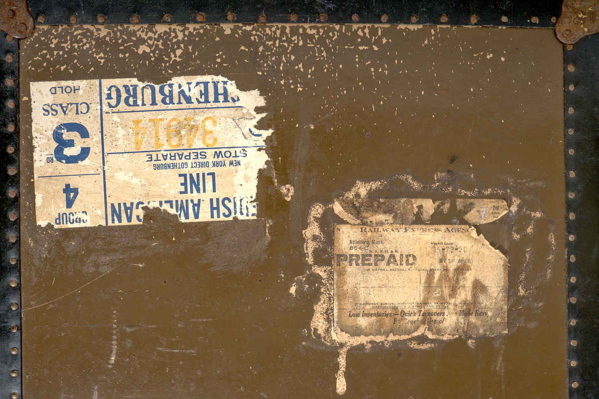 a) Rektangulär resekoffert med trästomme, utvändigt klädd i brunt papp och förstärkt med svartmålat konstläder. Kofferten är försedd med beslag och stötskydd av mässingspläterat stål. På vardera kortsida finns ett bärhandtag av kraftigt brunt läder. Koffertens framsida är försedd med låsanordningar bestående av två haspar samt i mitten ett ofullständigt lås, där låsdelen på kistans underdel saknas.

Invändigt har kofferten en uttagbar låda av papp, vilande på ribbor i kistans övre del. Insidan är klädd med grönt papper i geometriskt mönster. Bottendelen av lådan skyddas av ett smörpapper med blå prickar på vit botten. I kistans botten finns ett skyddande smörpapper med geometriska mönster i vitt, grönt och rött.

På vänster kortsida finns två stycken etiketter:

1. Swedish America Line.
2. Railway Express Agency (Attleboro, Massachusetts). Under denna etikett sitter fragment av en etikett i samma stil.

Se fotografi.

b) Pappersetikett med öljett, fäst i ståltråd: "Svenska.. Amerika. N: 936. Göteborgs Andra Bärarelag".