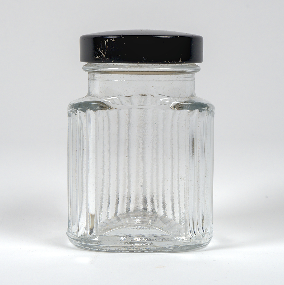 Opak glasburk med skruvlock av svart plast. Burken har en oval form och är dekorerad med vertikala räfflor i relief. På ena långsidan finns en plan yta, troligtvis avsedd för en etikett.

Burken är omärkt.