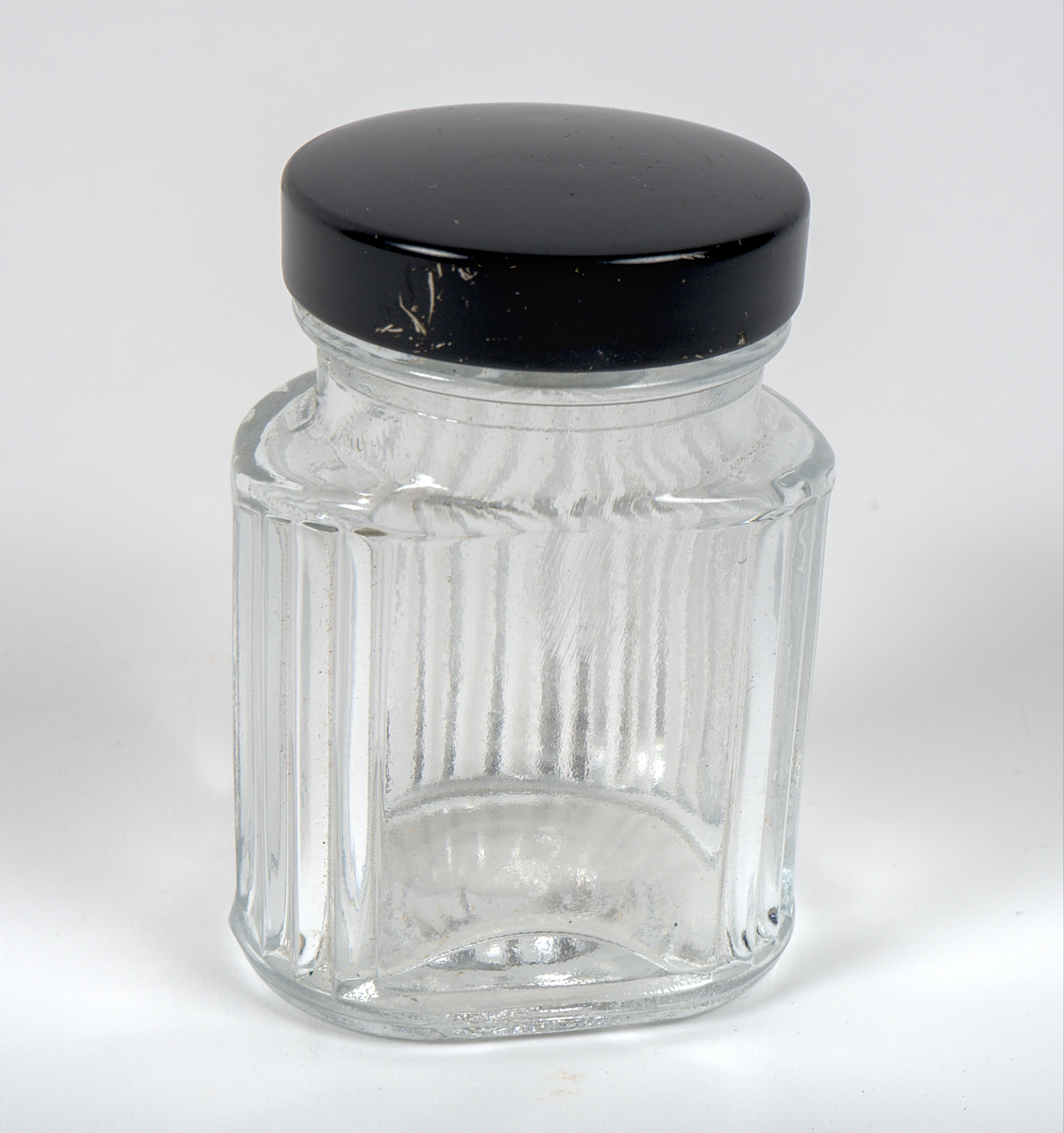 Opak glasburk med skruvlock av svart plast. Burken har en oval form och är dekorerad med vertikala räfflor i relief. På ena långsidan finns en plan yta, troligtvis avsedd för en etikett.

Burken är omärkt.