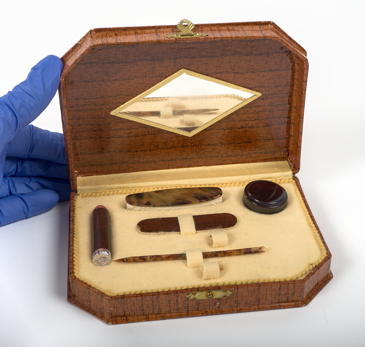 En uppsättning nagelvårdstillbehör med tillhörande etui.

a) Rektangulär ask av papp med yta i mahognyimitation. Framtill sitter en låsanordning i mässing, dekorerad med ett snäckformat beslag i art-deco stil. På insidan av locket finns en rombformad spegel, och på insidans botten en beige tyginsats kantad med ett flätat bomullsband.

b) Läppstift i cylindriskt format, inlindat i papper och aluminiumfolie. Längd: 50 mm.

c) Nagelpolerare med skaft av plast i sköldpaddsimitation. Nedre delen av mjukt material, klätt i läder. Längd: 62 mm. Bredd: 16 mm. Höjd 10 mm.

d) Nagelfil av papp med yta i mahognyimmitation. Sandpapper på undersidan. Längd 65 mm. Bredd 16 mm.

e) Nagelsticka av plast i sköldpaddsimitation. Längd: 90 mm.

f) Burk av plast. Tom på innehåll. Burken har troligtvis innehållet polermedel för nagelpoleraren.

De två tomrummen i asken visar att minst två delar saknas.