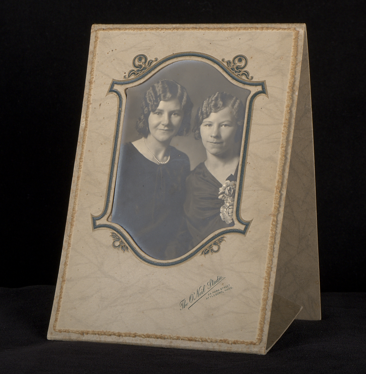 Fotografi monterat i etui av grå marmorerad papp. Etuiet är självstagande när det vecklas ut. Under fotografiet finns en stämpel: "The O'Neil Studio. 24 Park Street. Attleboro, Mass".

Fotografiet föreställer systrarna Edith (tv) och Sigrid (th) Torsson i finkläder.