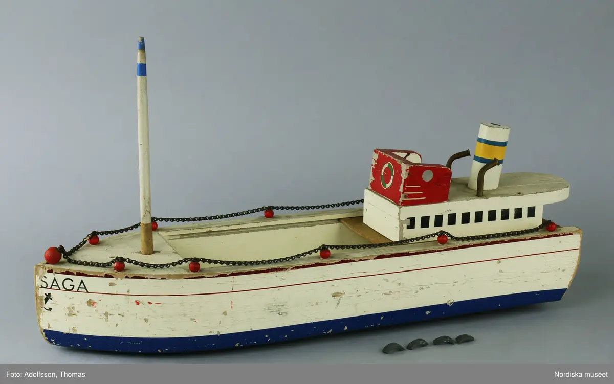 a) Lastbåten "SAGA" är av trä och är bemålad i vit, röd, blå gul, grön och svart färg. Färgen delvis avflagnad. Relingen och spikar av metall har rostat. b) Masten är löstagbar. På undersidan av båten finns fyra gummihjul och ett av c:1-4) gummihjulen har fallit sönder i bitar. Till båten hör troligen även ankaret NM.0335015 av svartmålad metall.