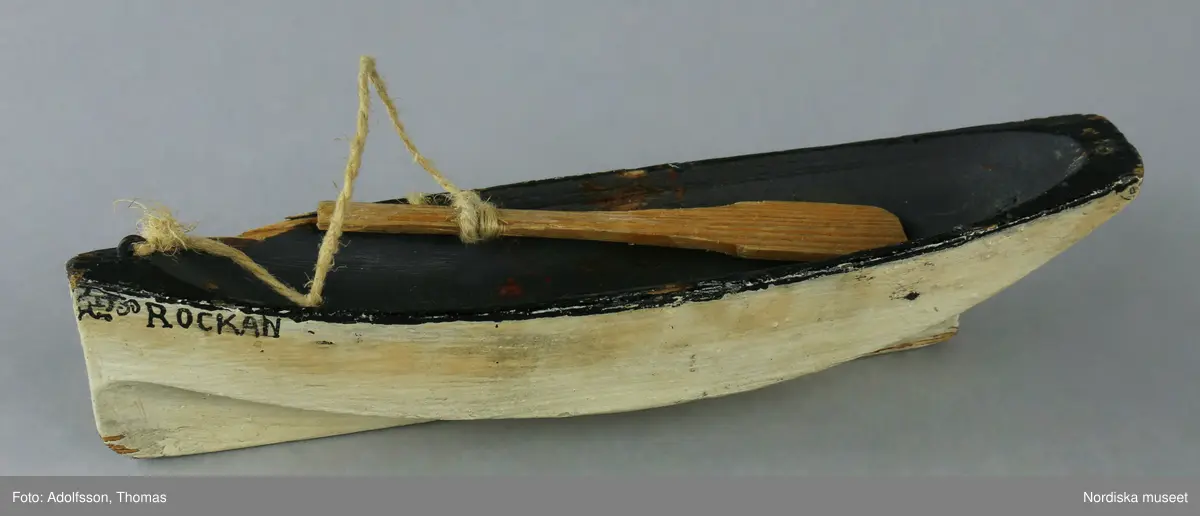 Leksaksbåten "Rockan" är gjord av trä och målad med vit färg på utsidan och svart inuti. En åra av naturfärgat trä sitter fast med ett snöre i en märla av metall.