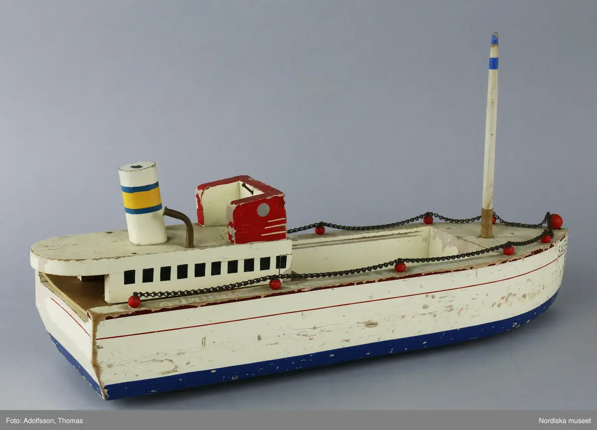 a) Lastbåten "SAGA" är av trä och är bemålad i vit, röd, blå gul, grön och svart färg. Färgen delvis avflagnad. Relingen och spikar av metall har rostat. b) Masten är löstagbar. På undersidan av båten finns fyra gummihjul och ett av c:1-4) gummihjulen har fallit sönder i bitar. Till båten hör troligen även ankaret NM.0335015 av svartmålad metall.