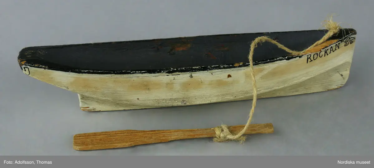 Leksaksbåten "Rockan" är gjord av trä och målad med vit färg på utsidan och svart inuti. En åra av naturfärgat trä sitter fast med ett snöre i en märla av metall.