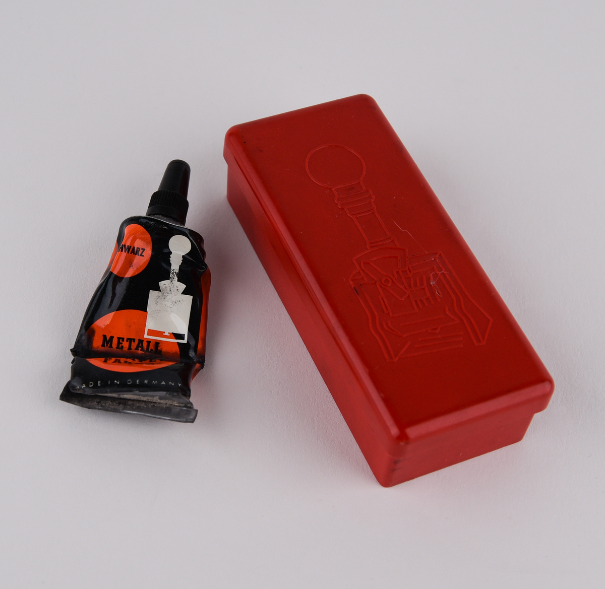 Stämpelfärg inlindad i papper i en röd ask av plast med en bild på en stämpel. Tysk text: "Schwartz, Metall Stempel Farbe". Består av tre delar: färgtub (Jvm23787-1-1), ask (Jvm23787-1-2) och lock till asken (Jvm23787-1-3).