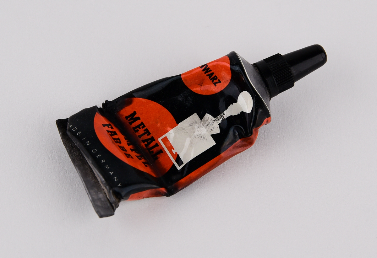 Stämpelfärg inlindad i papper i en röd ask av plast med en bild på en stämpel. Tysk text: "Schwartz, Metall Stempel Farbe". Består av tre delar: färgtub (Jvm23787-1-1), ask (Jvm23787-1-2) och lock till asken (Jvm23787-1-3).