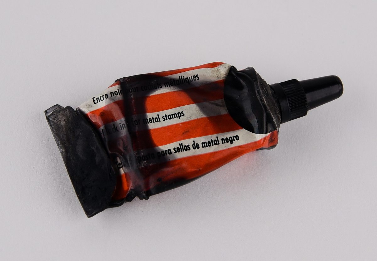 Stämpelfärg inlindad i papper i en röd ask av plast med en bild på en stämpel. Tysk text: "Schwartz, Metall Stempel Farbe". Består av tre delar: färgtub (Jvm23787-1-1), ask (Jvm23787-1-2) och lock till asken (Jvm23787-1-3).