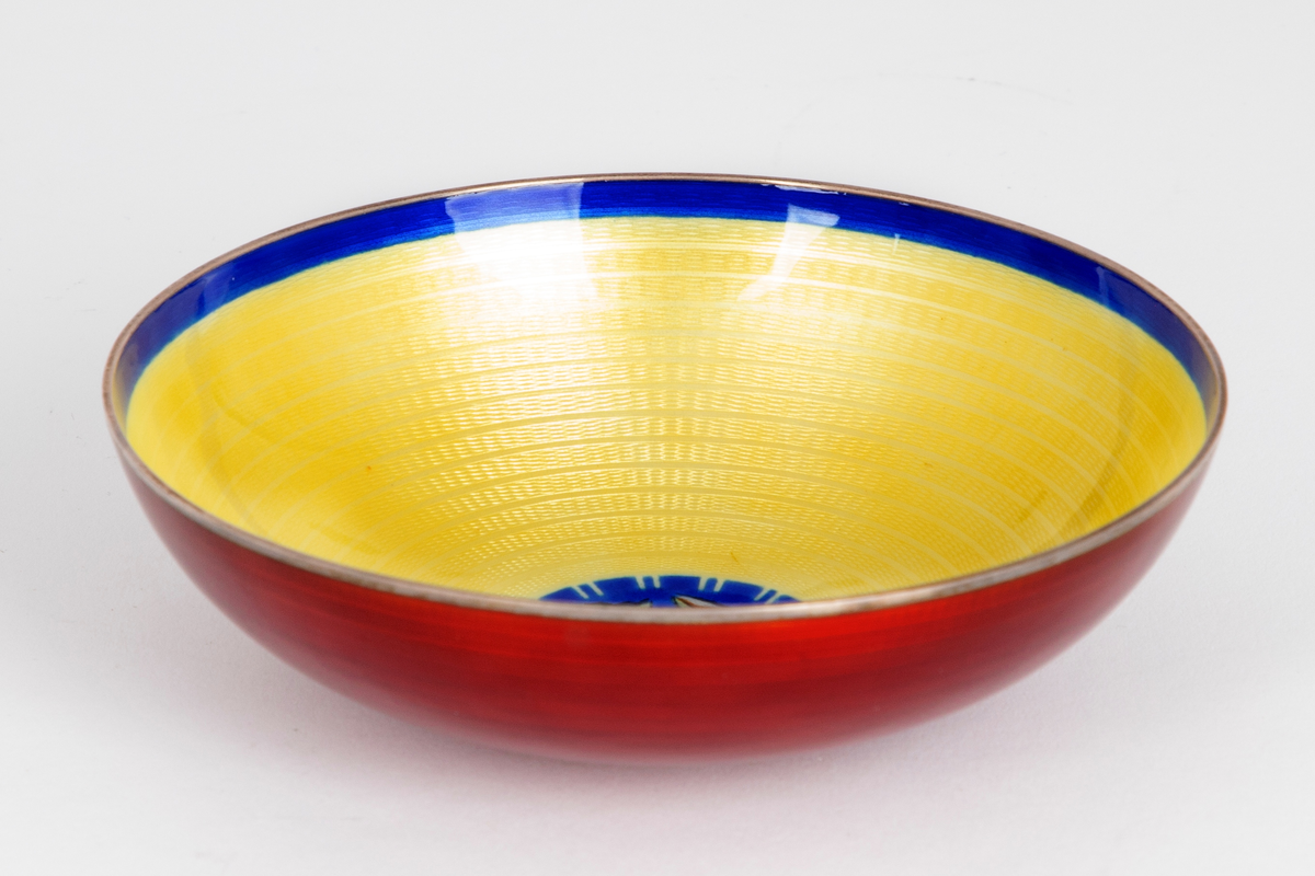 Skål i emaljert sølv. Konkav form med lav fotring. Utvendig rød emalj på guillochert bunn, innvendig gul og blå emalje samt et fiskemotiv på guillochert bunn.