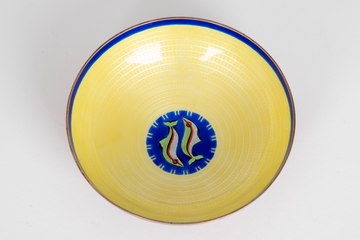 Skål i emaljert sølv. Konkav form med lav fotring. Utvendig rød emalj på guillochert bunn, innvendig gul og blå emalje samt et fiskemotiv på guillochert bunn.