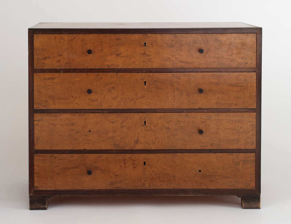 Rektangulær, rettlinjet kommode i flammebjørkfinér på tre, med en ramme i mørkere finér. Kommoden har fire skuffer, på hver skuff et midtstilt nøkkelhull og to små, runde knopper som håndtak, en på hver side av nøkkelhullet. To av knoppene mangler. Kommoden står på lave, firkantete fotstykker integrert i rammen.