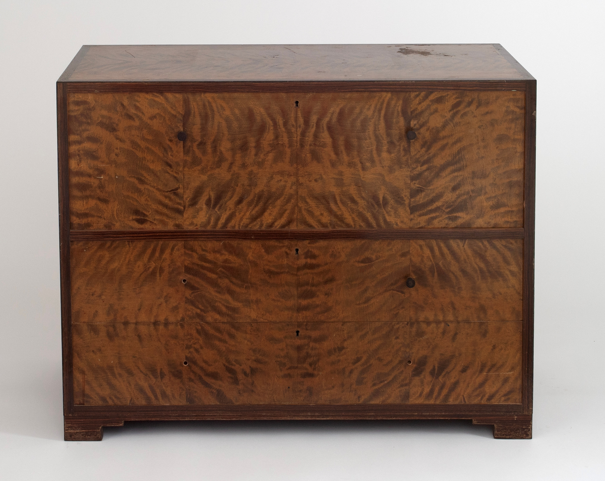 Rektangulær, rettlinjet kommode i flammebjørkfinér på tre, med en ramme i mørkere finér. Kommoden har tre skuffer, øverst en dyp skuff og nederst to mindre skuffer. På hver skuff et midtstilt nøkkelhull og to små, runde knopper som håndtak, en på hver side av nøkkelhullet. Tre av knoppene mangler. Kommoden står på lave, firkantete fotstykker integrert i rammen.