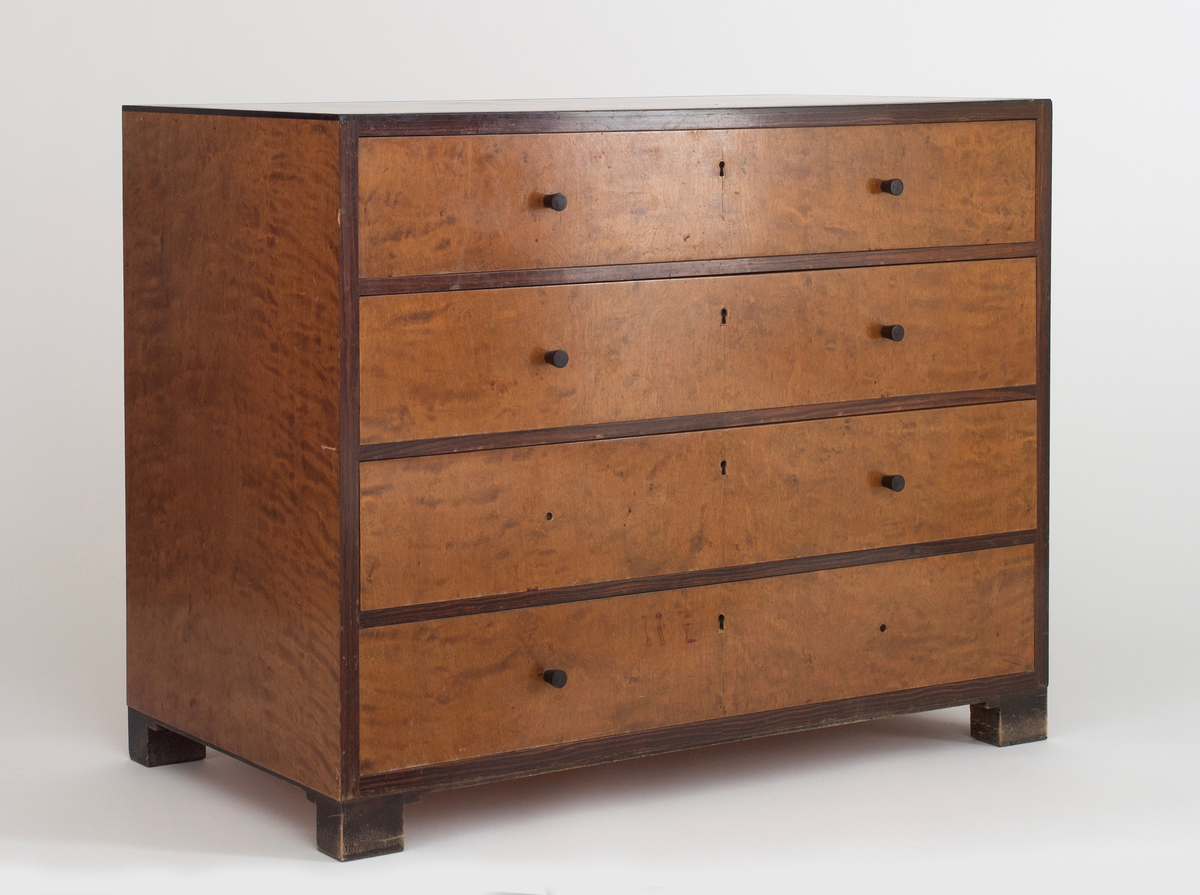 Rektangulær, rettlinjet kommode i flammebjørkfinér på tre, med en ramme i mørkere finér. Kommoden har fire skuffer, på hver skuff et midtstilt nøkkelhull og to små, runde knopper som håndtak, en på hver side av nøkkelhullet. To av knoppene mangler. Kommoden står på lave, firkantete fotstykker integrert i rammen.