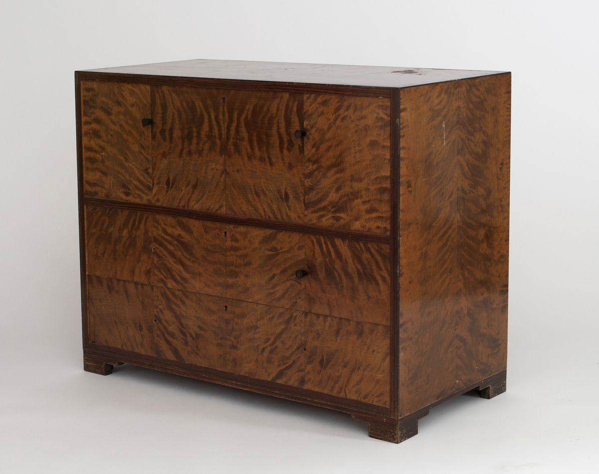 Rektangulær, rettlinjet kommode i flammebjørkfinér på tre, med en ramme i mørkere finér. Kommoden har tre skuffer, øverst en dyp skuff og nederst to mindre skuffer. På hver skuff et midtstilt nøkkelhull og to små, runde knopper som håndtak, en på hver side av nøkkelhullet. Tre av knoppene mangler. Kommoden står på lave, firkantete fotstykker integrert i rammen.