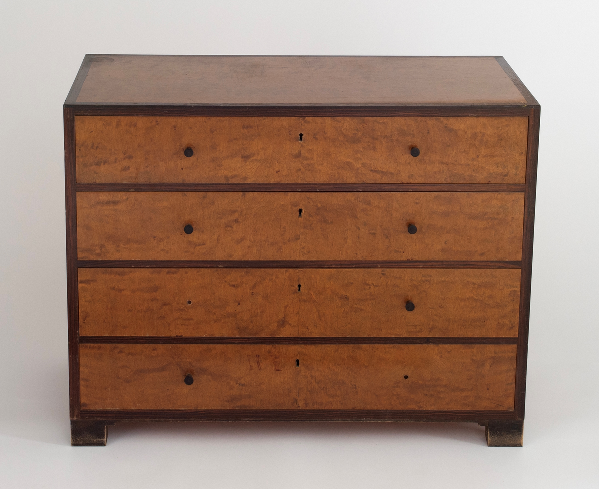 Rektangulær, rettlinjet kommode i flammebjørkfinér på tre, med en ramme i mørkere finér. Kommoden har fire skuffer, på hver skuff et midtstilt nøkkelhull og to små, runde knopper som håndtak, en på hver side av nøkkelhullet. To av knoppene mangler. Kommoden står på lave, firkantete fotstykker integrert i rammen.