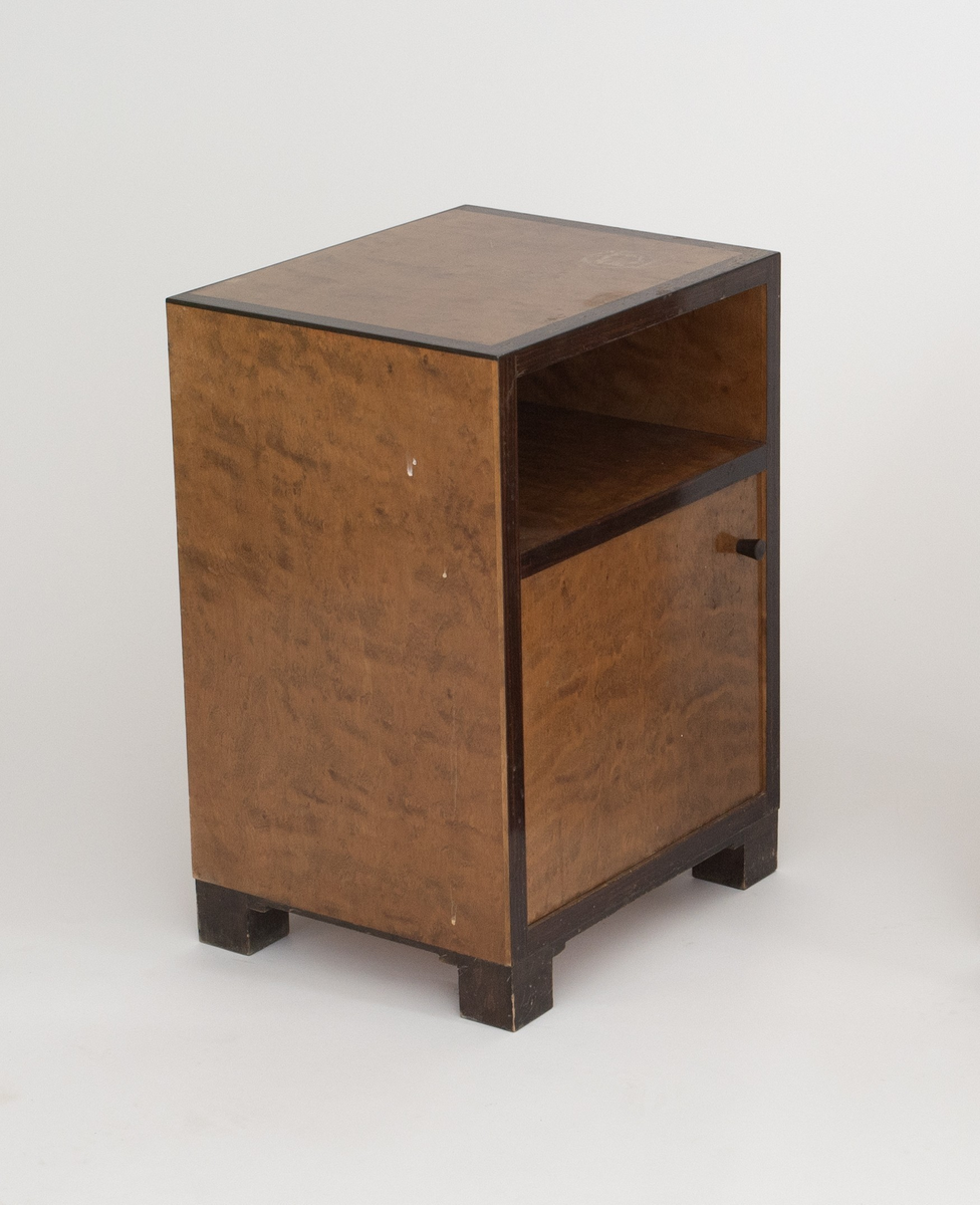 Rektangulært, rettlinjet nattbord i flammebjørkfinér på tre, med en ramme i mørkere finér. Nattbordet har en hylle samt et skap, på skapdøren en liten, rund knopp i øverste høyre hjørne som håndtak. Står på lave, firkantete fotstykker integrert i rammen.