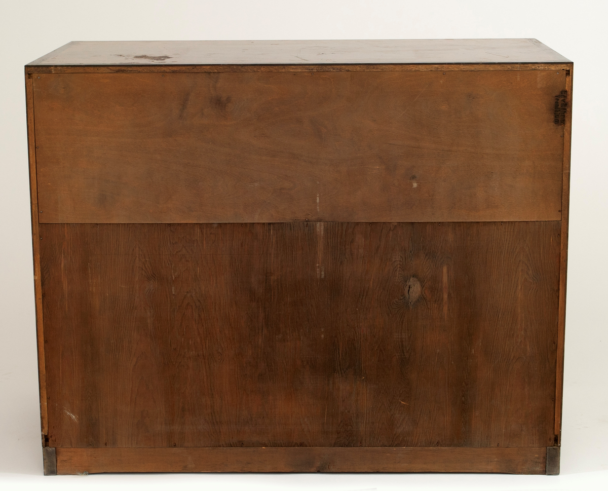 Rektangulær, rettlinjet kommode i flammebjørkfinér på tre, med en ramme i mørkere finér. Kommoden har tre skuffer, øverst en dyp skuff og nederst to mindre skuffer. På hver skuff et midtstilt nøkkelhull og to små, runde knopper som håndtak, en på hver side av nøkkelhullet. Tre av knoppene mangler. Kommoden står på lave, firkantete fotstykker integrert i rammen.