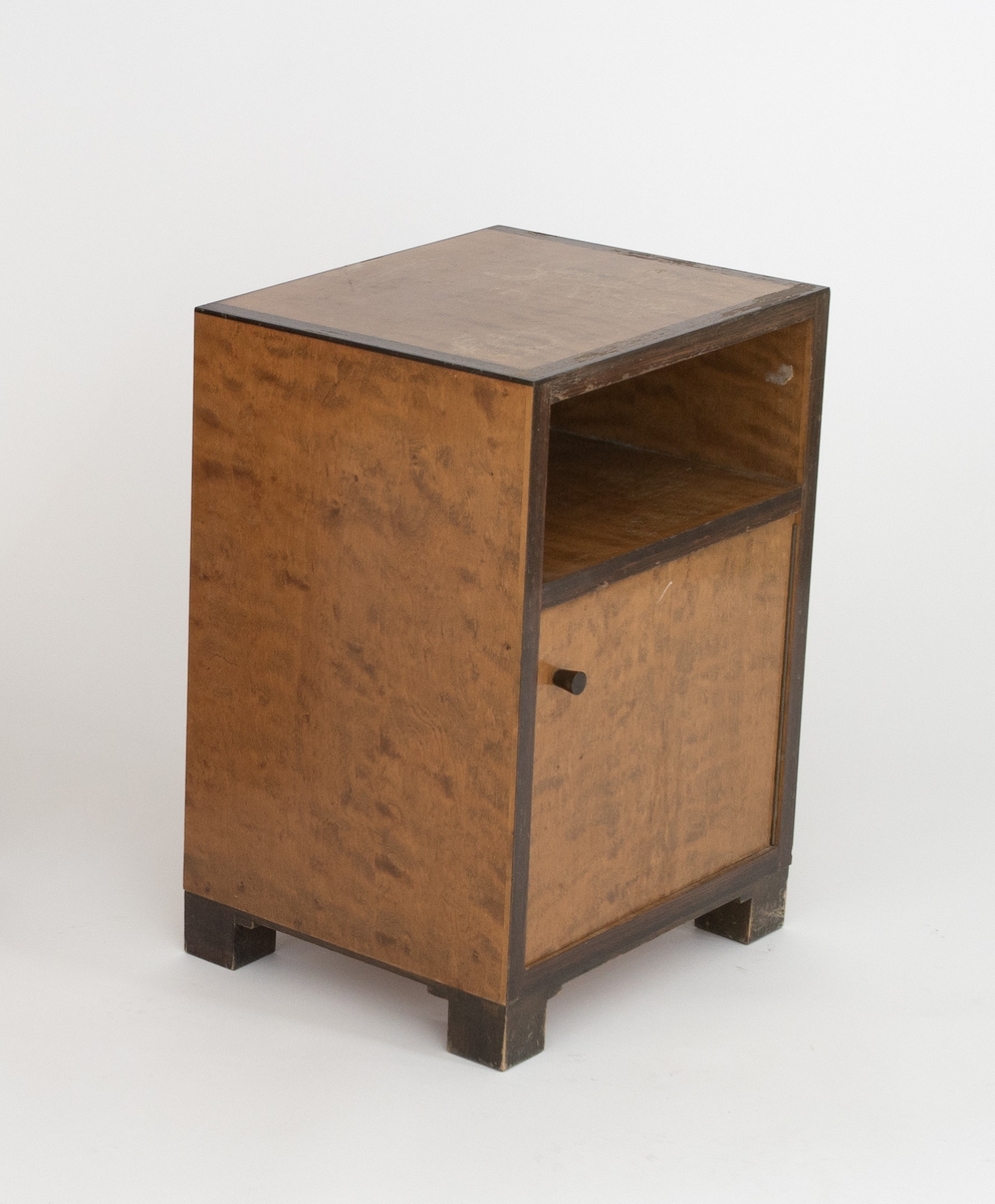 Rektangulært, rettlinjet nattbord i flammebjørkfinér på tre, med en ramme i mørkere finér. Nattbordet har en hylle samt et skap, på skapdøren en liten, rund knopp i øverste venstre hjørne som håndtak. Står på lave, firkantete fotstykker integrert i rammen.