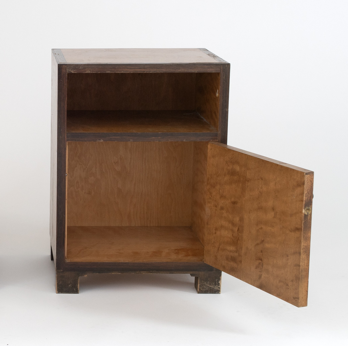 Rektangulært, rettlinjet nattbord i flammebjørkfinér på tre, med en ramme i mørkere finér. Nattbordet har en hylle samt et skap, på skapdøren en liten, rund knopp i øverste venstre hjørne som håndtak. Står på lave, firkantete fotstykker integrert i rammen.