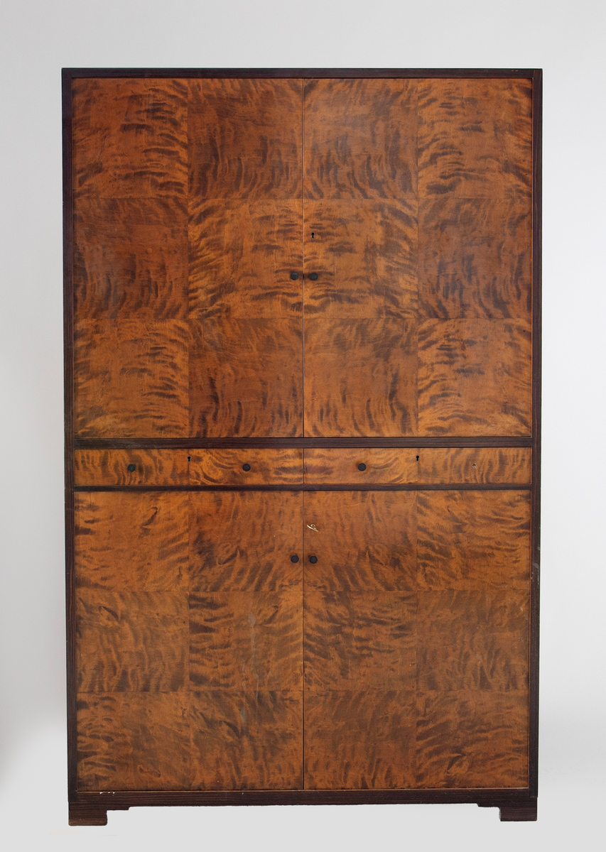 Rektangulært, rettlinjet skap i flammebjørkfinér på tre, med en ramme i mørkere finér. Øverst et skap, i midten to skuffer ved siden av hverandre og nederst et skap. Skapene har dobbeltdører med en liten, rund knopp som håndtak på hver dør. Nøkkelhull på de høyre skapdørene. Skapene er innredet med tre hyller. På hver skuff et midtstilt nøkkelhull og to små, runde knopper som håndtak, en på hver side av nøkkelhullet. Skapet står på lave, firkantete fotstykker integrert i rammen.