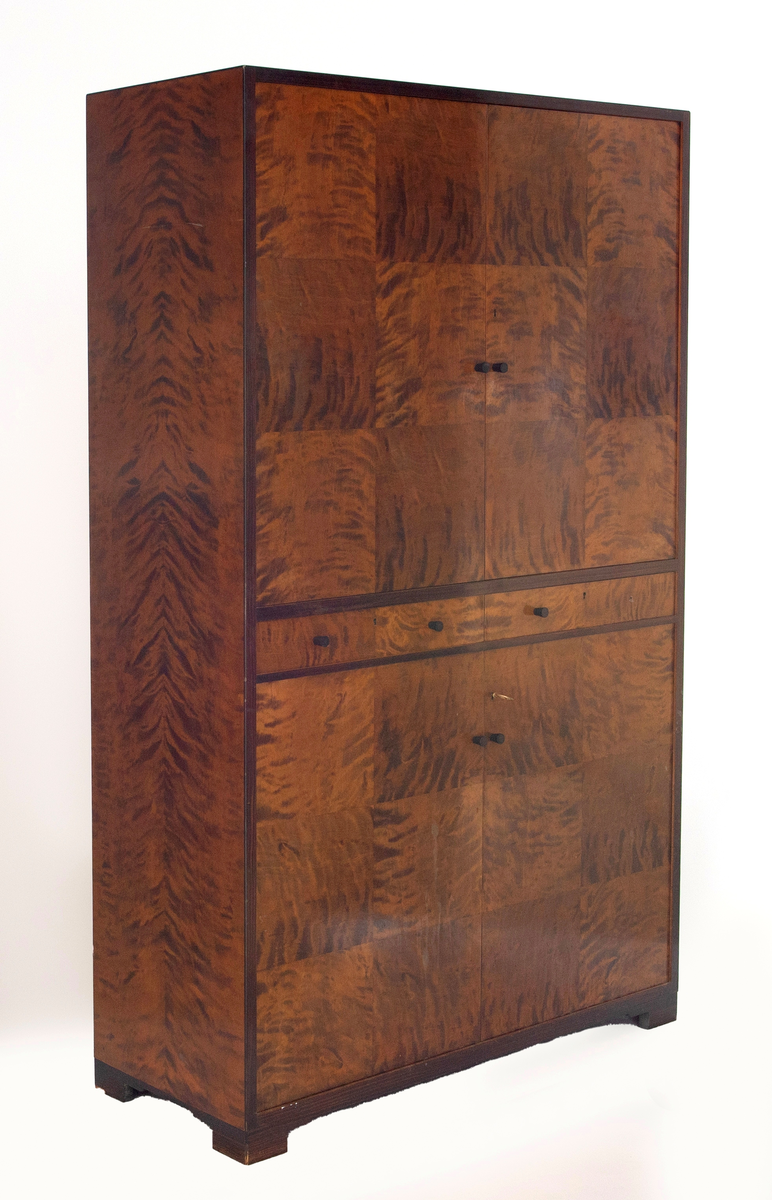 Rektangulært, rettlinjet skap i flammebjørkfinér på tre, med en ramme i mørkere finér. Øverst et skap, i midten to skuffer ved siden av hverandre og nederst et skap. Skapene har dobbeltdører med en liten, rund knopp som håndtak på hver dør. Nøkkelhull på de høyre skapdørene. Skapene er innredet med tre hyller. På hver skuff et midtstilt nøkkelhull og to små, runde knopper som håndtak, en på hver side av nøkkelhullet. Skapet står på lave, firkantete fotstykker integrert i rammen.