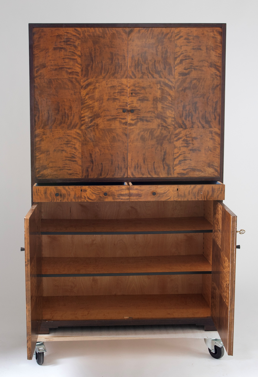 Rektangulært, rettlinjet skap i flammebjørkfinér på tre, med en ramme i mørkere finér. Øverst et skap, i midten to skuffer ved siden av hverandre og nederst et skap. Skapene har dobbeltdører med en liten, rund knopp som håndtak på hver dør. Nøkkelhull på de høyre skapdørene. Skapene er innredet med tre hyller. På hver skuff et midtstilt nøkkelhull og to små, runde knopper som håndtak, en på hver side av nøkkelhullet. Skapet står på lave, firkantete fotstykker integrert i rammen.