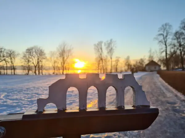 Porten inn til Storhamarstuene er dekorert med en stilisert utskjæring av domkirkeruinen, her i fin silhuett mot solnedgang over snødekt landskap og med Storhamarstuene i bakgrunnen.
